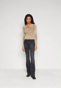 Moteris vilkinti prigludusį smėlio spalvos ilgų rankovių marškinį su volangomis ir juodus pla�čius džinsus, stovinti prieš paprastą baltą foną.