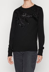 Schwarzer Pullover mit einem strukturierten Design aus schwarzen Pailletten in Wellenform, geripptem Halsausschnitt und Bündchen. Weicher Stoff und lockere Passform.