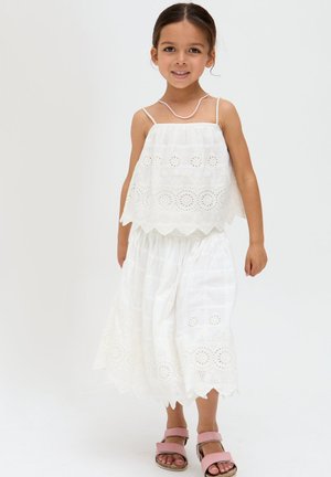 Junges Mädchen trägt ein weißes, besticktes ärmelloses Oberteil und passenden Rock, pinke Sandalen und eine pinke Perlenkette, steht und lächelt.