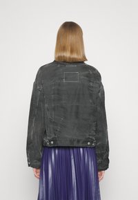 Svart denimjacka med en lätt sliten textur, som har en fyrkantig patch på övre ryggen och tryckknappsstängning vid nederkanten.