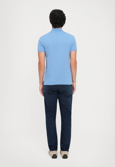 Man met krullend haar, gekleed in een lichtblauw polo-shirt, donkerblauwe jeans en beige schoenen, staat met zijn rug naar de camera tegen een plain achtergrond.