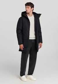 Parka nera imbottita con cappuccio, chiusura con zip e tasche frontali. Indossata sopra un maglione beige e abbinata a jeans scuri e sneaker bianche.