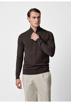 Uomo che indossa un maglione marrone scuro con cerniera e pantaloni beige, che regola la cerniera, in piedi davanti a uno sfondo chiaro e uniforme.