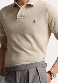 Polo shirt beige con tejido de punto texturizado, que presenta una tapeta de tres botones y un pequeño logo marrón en el pecho. Pantalones grises con pliegues.