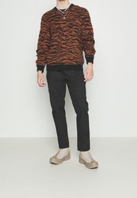 Orange and black patterned sweater with a crew neckline, paired with black pants and light beige lace-up shoes. Minimal accessories visible.