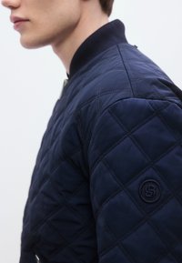 Μπλε ναυτικό μπουφάν bomber με ριμπ γιακά, διαγώνιο ράψιμο και κυκλικό λογότυπο στον αριστερό ώμο.