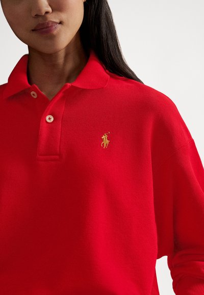 Rød poloshirt laget av strikket stoff med en knappet krage og en gullbrodert logo på venstre bryst.