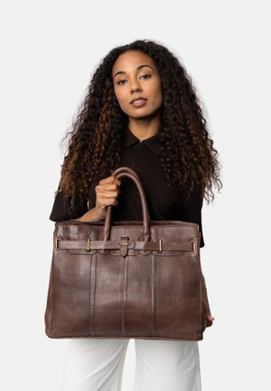 Vrouw met lang krullend haar houdt een grote bruine leren tote bag vast voor een witte achtergrond, gekleed in een zwart top en witte broek.