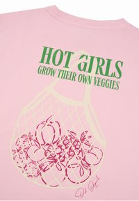 Ροζ πουκάμισο με πράσινο κείμενο «HOT GIRLS GROW THEIR OWN VEGGIES» πάνω από μια εκρού δίχτυνη τσάντα γεμάτη με εικονογραφημένα κόκκινα φρούτα και λαχανικά.