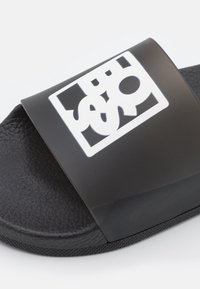 Sandale noire à enfiler avec semelle texturée et large bride avec un motif logo graphique blanc.