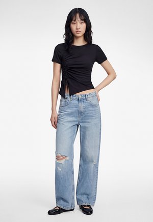 Jonge vrouw staat, gekleed in een zwart gerimpeld kortarmig topje, lichtblauwe gescheurde jeans met wijde pijpen en zwarte platte schoenen, tegen een witte achtergrond.