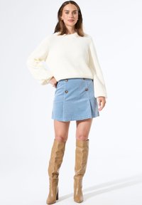 Vrouw staand met hand op heup, gekleed in een crèmekleurige gebreide trui, blauwe button-down minirok en tan kleurige kniehoge laarzen op een witte achtergrond.