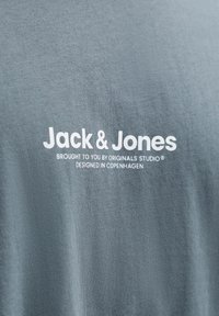 Γκρι ύφασμα με λευκό κείμενο "Jack & Jones, φέρνει σε εσάς το Originals Studio, σχεδιασμένο στην Κοπεγχάγη" τυπωμένο πάνω του.