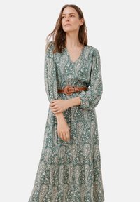 Langes, grünes Paisleymuster-Kleid mit tiefem V-Ausschnitt und langen Ärmeln, tailliert mit einem braunen Gürtel mit einer runden Schnalle.