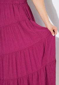 Fuchsia-tägliches Kleid aus leichtem, strukturiertem Stoff. Verfügt über fließende Lagen und Rüschen, mit einer Hand, die den Saum hält.