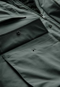 Chaqueta verde oscuro con múltiples bolsillos, que presenta una textura suave y mate, con broches de presión negros para un cierre seguro. Detalle de cremallera visible.