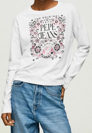 Henkilö pukeutuneena valkoiseen pitkähihainen paita, jossa musta ja pinkki kukkakuosi ja teksti "Pepe Jeans London 1973", yhdistettynä sinisiin farkkuihin.