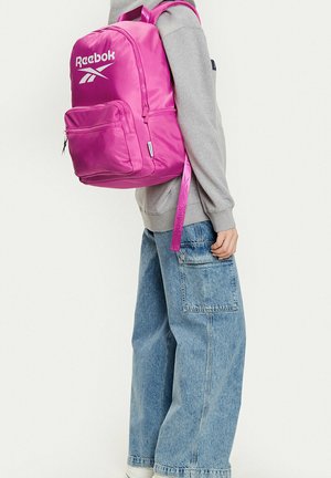 Osoba ubrana w szarą bluzę i szerokie niebieskie dżinsy z jasnoróżowym plecakiem Reebok na jednym ramieniu na tle białego tła.