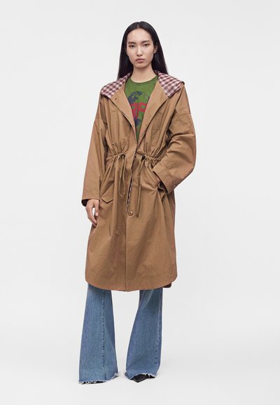Femme portant un manteau marron oversize avec une taille à cordon, une doublure de capuche à carreaux roses, un t-shirt vert à motif graphique et un jean bleu évasé.