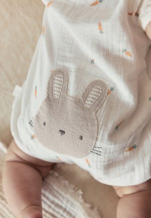 Vauva, joka käyttää valkoisia vaatteita, joissa on pieniä porkkanaprinttejä ja ommeltu beige pupun kasvot edessä, istuu teksturoidulla pinnalla.
