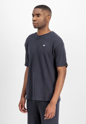 Junger Mann, der ein dunkelgraues, kurzärmeliges, strukturiertes Hemd und passende Hosen trägt, vor einem schlichten weißen Hintergrund stehend.