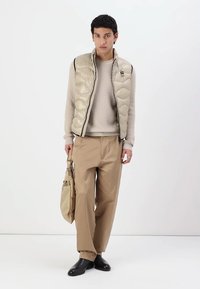 Gilet imbottito beige con chiusura a zip su un maglione beige a trama; abbinato a pantaloni crema e scarpe nere, mentre tiene una borsa a tracolla color mogano.