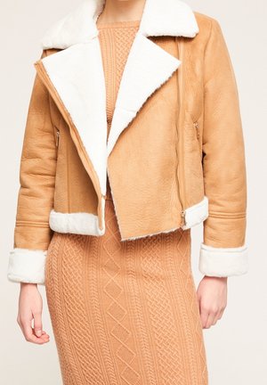 Tan shearling jacket with white fur trim worn over a textured camel cable-knit dress, with visible zippers and folded cuffs.