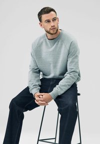 Grå sweatshirt med tekstureret finish, rund hals og ribbede manchetter; parret med mørke denimjeans med kontrastsying. Sidder på en skammel.
