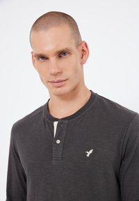 Long-sleeve henley shirt in dark gray, featuring a buttoned neckline and a cream panel. Notable white embroidered bird logo on the chest.