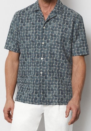 Muž v krátkém rukávu s košilí na knoflíky s modrým geometrickým vzorem krychlí a bílými kalhotami stojící před jednobarevným pozadím.