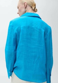 Camicia turchese a maniche lunghe realizzata in tessuto leggero, con colletto classico, chiusure a bottoni ai polsini e orlo leggermente curvato.
