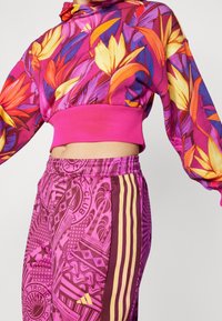 Tajt vy av en person som bär en färgstark blommig hoodie i lila, orange och gula nyanser samt rosa träningsbyxor med grafiska mönster och gula ränder.