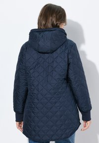 Giacca trapuntata blu navy con cappuccio, polsini elasticizzati e un retro leggermente più lungo, caratterizzata da una texture a motivo diamante.