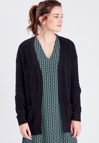 Gilet en maille noir avec une ouverture sur le devant, des poignets côtelés et des poches latérales, superposé sur une robe à motifs floraux verts.
