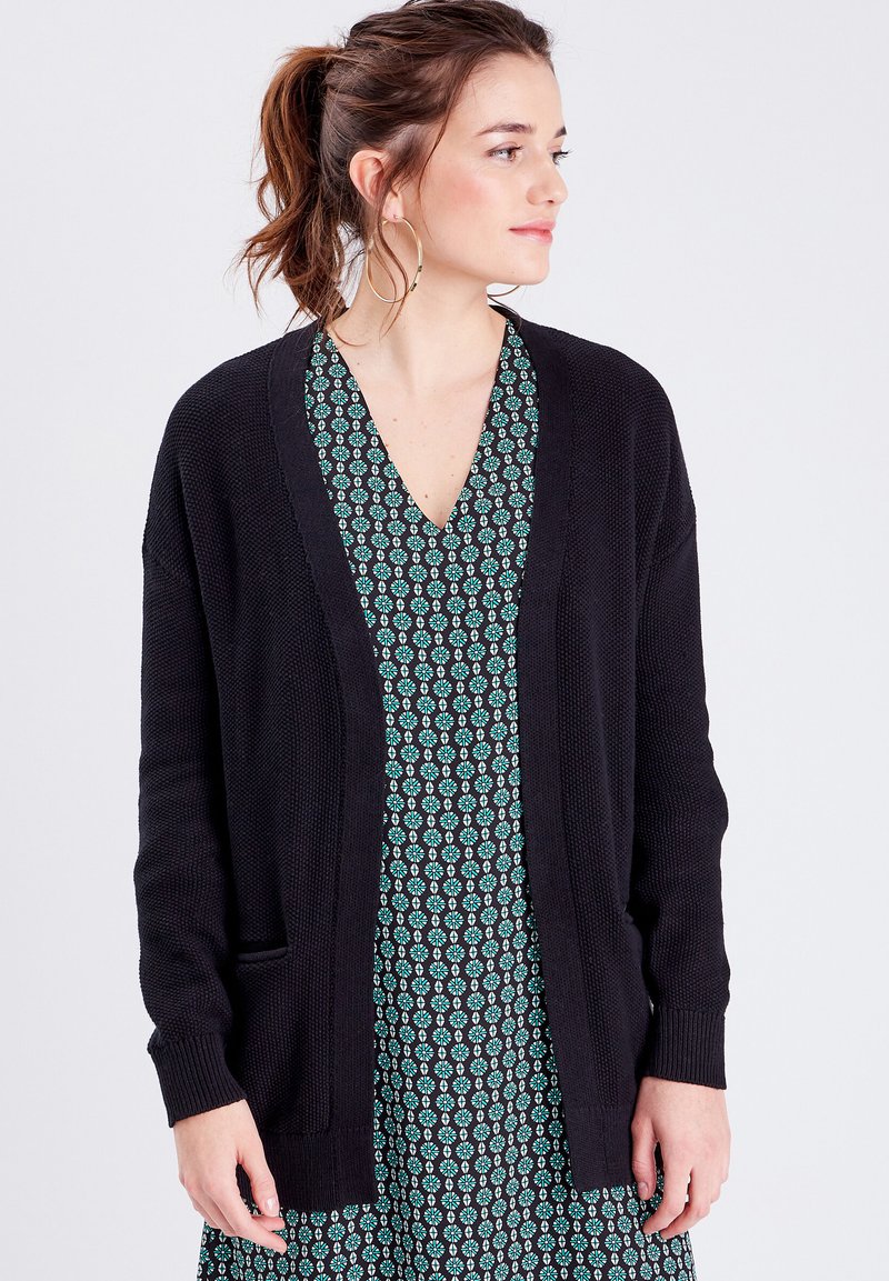 Gilet en maille noir avec une ouverture sur le devant, des poignets côtelés et des poches latérales, superposé sur une robe à motifs floraux verts.