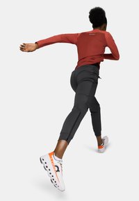 Un athlète portant un haut à manches longues rouge et un pantalon noir ajusté, courant avec des baskets blanches et orange, présentant des accents réfléchissants.