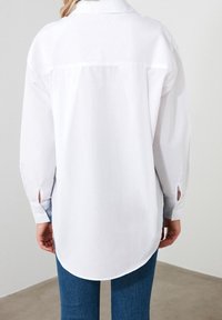 Camicia bianca con yoke posteriore, maniche lunghe e fessure laterali. Realizzata in tessuto liscio, presenta polsini con bottone e orlo arrotondato.