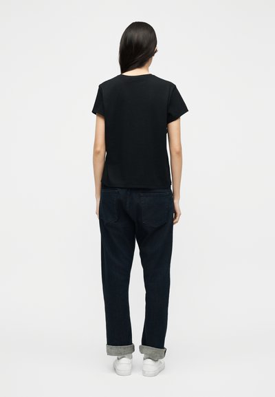 Γυναίκα με μακριά σκούρα μαλλιά, φορώντας μαύρο κοντομάνικο πουκάμισο, σκουρόχρωμα τζιν με ρεβέρ και λευκά αθλητικά παπούτσια, στέκεται πλάτη σε απλό φόντο.