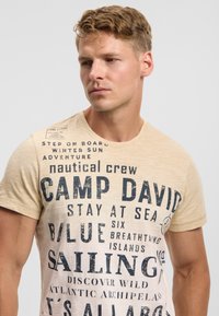 Light beige short-sleeve t-shirt featuring dark navy text, phrases, and a shoulder tag. The fabric has a soft, slightly textured appearance.