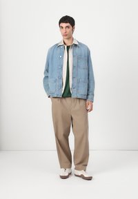 Denim jacket in light blue, features chest pockets, layered over a green shirt and beige trousers, completed with white and brown sneakers.