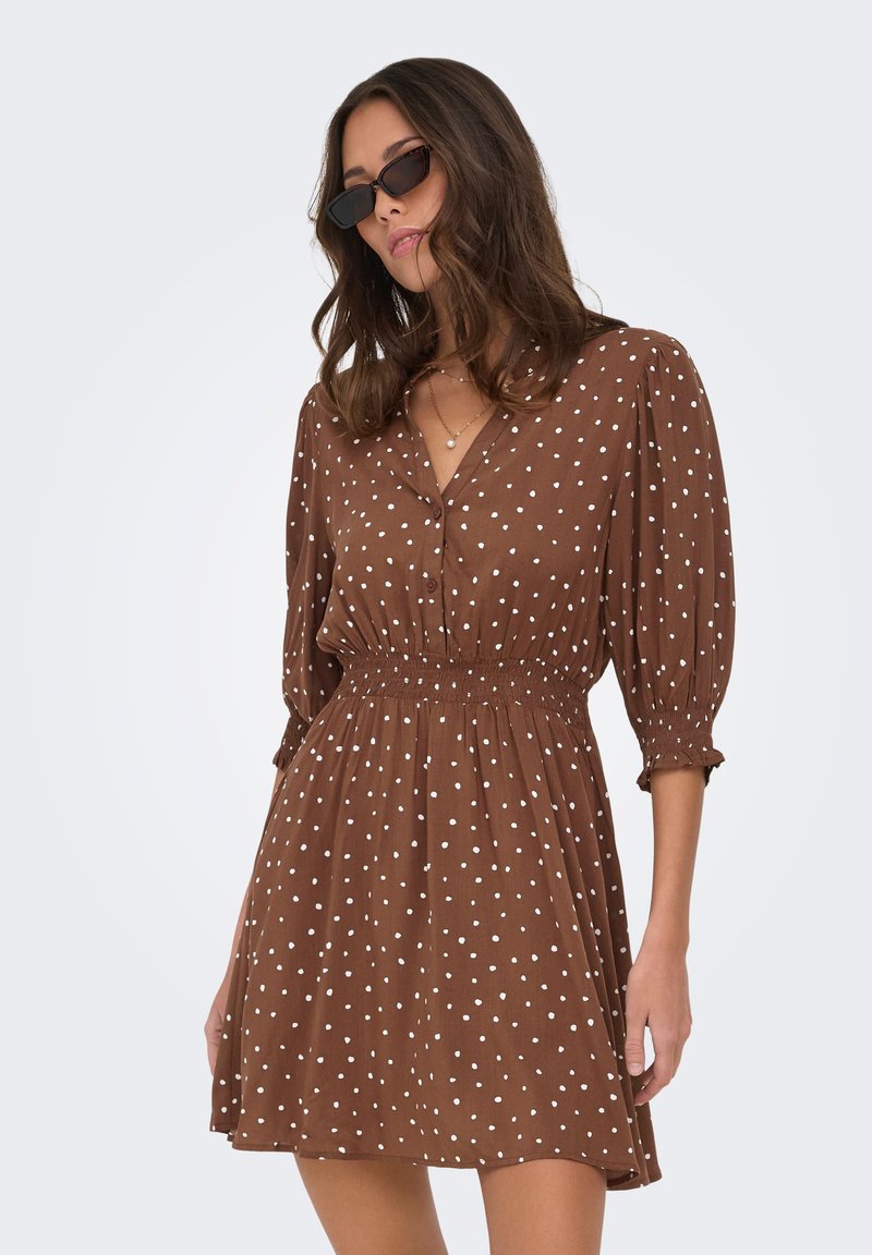 Femme portant une robe marron à pois blancs, taille smockée et manches trois quarts, accessoirisée avec des lunettes de soleil noires et des colliers superposés.