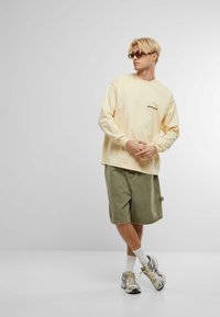 Chemise à manches longues jaune clair avec du texte dans le dos, associée à un short vert olive. Le modèle porte des chaussettes blanches et des baskets multicolores.