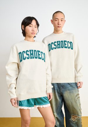 Deux personnes portent des sweats crème avec "DCSHOECO" en texte teal. L'un porte un short satin teal avec des bordures en dentelle ; l'autre a un jean déchiré.