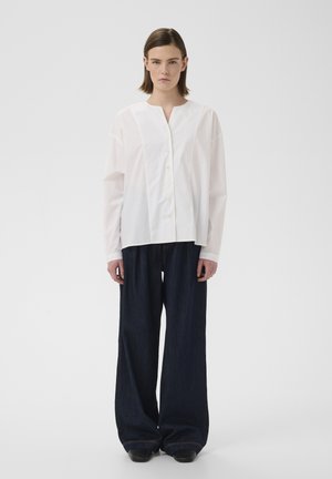 Vrouw staat frontaal, draagt een losse witte blouse met knopen en een wijde donkerblauwe spijkerbroek tegen een effen witte achtergrond.