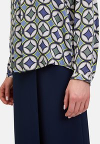 Langärmlige Bluse mit geometrischem Muster in Blau, Grün und Weiß, mit einem fließenden Design und abgerundetem Saum. Trägt sie über dunklen Marineblauen Hosen.