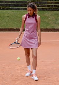 Ružové tenisové šaty s vínovým golierom, vyrobené z hladkej látky. Na nohách má biele ponožky a tenisové topánky s ružovými akcentmi, pričom drží raketu.