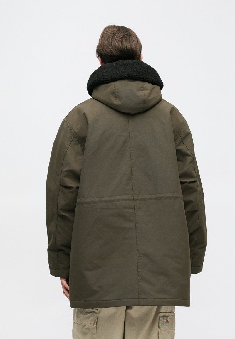 Cappotto lungo verde oliva con un collo nero e morbido. Presenta un cappuccio e un tessuto texturizzato, con cuciture visibili sul retro.