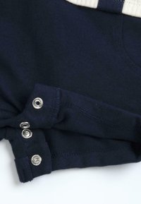 Tissu bleu marine avec bordure blanche, présentant trois boutons-pression argentés partiellement ouverts sur le bord d'un vêtement, sur fond blanc.