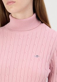 Pull rose tricoté avec un col montant, présentant un motif torsadé et un petit logo brodé sur la poitrine. Texture douce et coupe ajustée.
