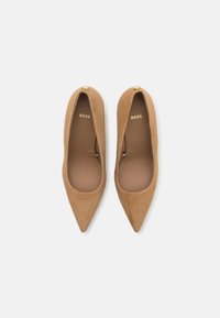 Beige suède, hegyes orrú pump, sima felülettel. Elülső részén egy kis arany dísz és a belsejében egy berakott márkajelzés található.